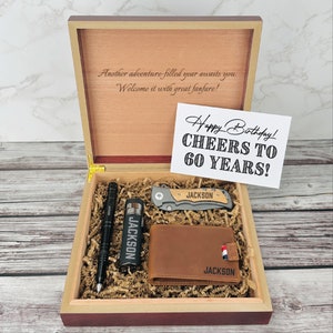 May include: A wooden gift box with a brown leather wallet, a pocket knife, a pen, and a lighter. The box is open and the items are arranged inside. The box has a personalized message that says "Another adventure filled year awaits you. Welcome it with great fanfare!" and a card that says "Happy Birthday! Cheers to 60 years!"