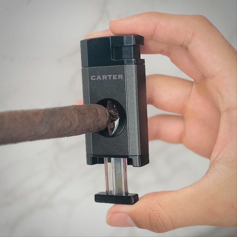 May include: A black and silver cigar cutter with the word "CARTER" printed on the side. A cigar is being cut by the cutter.