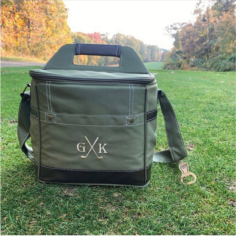 May include: A green canvas cooler bag with a black leather trim and a brown leather handle. The bag has a zipper closure and a monogrammed design with the initials "G.X.K." and crossed golf clubs. A gold bottle opener is attached to the bag.