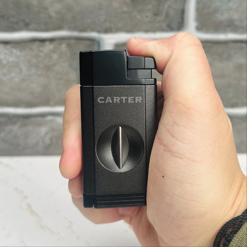 May include: A black cigar cutter with the word "CARTER" printed on the front. The cutter has a round, silver blade.