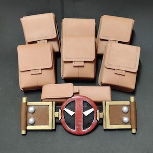 May include: A brown leather belt with a red and black Deadpool logo buckle. The belt has six brown leather pouches attached to it.