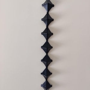 Könnte beinhalten: Eine vertikale, schwarze, geometrische Kette aus rautenförmigen Gliedern hängt vor einer neutralen Wand. Jedes Glied ist ein dunkles, mattes Schwarz, und die Kette scheint aus Metall oder einem ähnlichen Material zu bestehen.