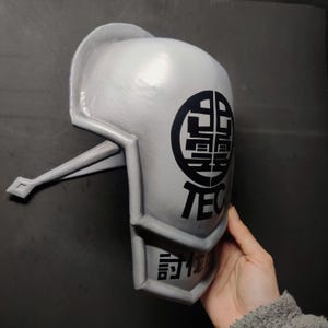 Könnte beinhalten: Ein grauer Plastikhelm mit schwarzer Beschriftung und einem kreisförmigen Design. Der Helm hat einen erhöhten Rand um den Rand und einen Riemen an der Seite. Der Text auf dem Helm lautet "TEC" und "言语行动".