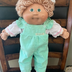May include: A Cabbage Patch Kids doll with light brown yarn hair, green eyes, and a dimple on the left cheek. The doll wears a white short-sleeved shirt with lace trim and pink stitching, paired with mint green corduroy overalls and white shoes.