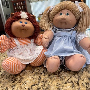 May include: Two dolls with yarn hair and soft fabric bodies. The left doll has brown yarn hair, blue eyes, and wears an orange and white striped outfit with a white pinafore. The right doll has blonde yarn hair, green eyes, and a blue gingham dress.