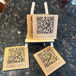 May include: Four wooden coasters with a black and white QR code and the text "Menu Items" printed on each. The coasters are stacked on a wooden easel.