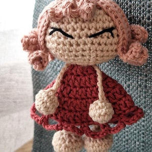 May include: A crocheted doll with pink hair, a red dress, and white pom-poms. The doll has a simple face with black eyes and a closed mouth.