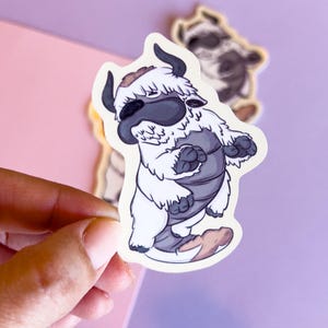 May include: A sticker featuring a cartoon bison-like creature with white fur, gray body, and black horns. The sticker is held by a hand against a purple background, with a second sticker of the same design visible in the background. The creature has a friendly expression.