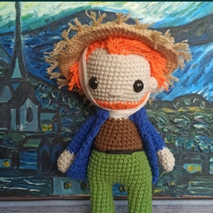 Könnte beinhalten: Häkelpuppe mit orangefarbenem Haar, die einen Strohhut und eine blaue Jacke trägt. Die Puppe steht vor einem Gemälde einer sternenklaren Nacht.