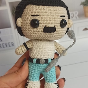 Puede incluir: Muñeco de ganchillo de un hombre con cabello negro y bigote, que lleva una camiseta sin mangas blanca, pantalones azules y zapatos blancos y negros. El muñeco sostiene un micrófono gris. El muñeco tiene ojos negros y lleva un cinturón negro.