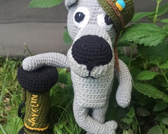 Puede incluir: Una figura de crochet hecha a mano de un perro gris que lleva un sombrero verde con un emblema azul y amarillo. El perro sostiene un objeto de crochet negro y verde con la palabra "JAVELIN" en amarillo. El perro está en la hierba.