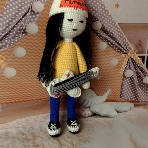 May include: Crocheted doll with long black hair, wearing a yellow shirt, blue trousers, and black shoes. The doll holds a guitar and wears a white beanie with the word "FUNK" in orange. The doll is in front of a tent.