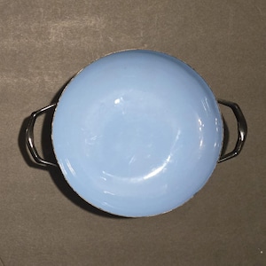 May include: A light blue enamelware bowl with two silver-colored handles. The bowl has a rounded shape and a smooth, glossy finish. The handles are curved and attached to the sides of the bowl. The bowl is empty.