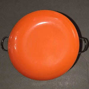 May include: A round, orange enamel pan with two black handles. The pan has a glossy finish and appears to be vintage. The handles are curved and attached to the sides of the pan. This cookware is suitable for cooking.