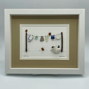 May include: Framed artwork depicting a clothesline with various sea glass and shell pieces hanging as laundry. The artwork includes small bird figures and the text "Wash Day". The frame is white.