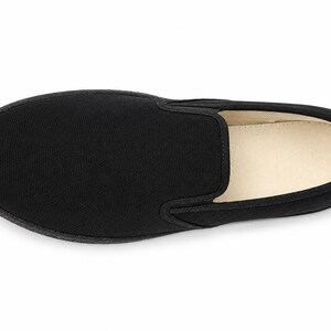 May include: A single black slip-on shoe, viewed from above, showcasing a minimalist design. The shoe features a solid black canvas upper, a rounded toe, and a contrasting off-white interior. The sole is black and appears to be made of rubber.