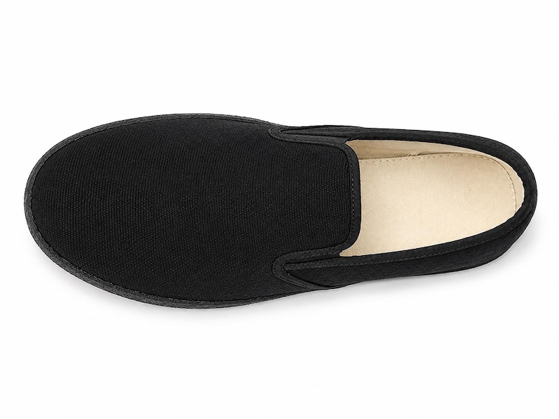 May include: A single black slip-on shoe, viewed from above, showcasing a minimalist design. The shoe features a solid black canvas upper, a rounded toe, and a contrasting off-white interior. The sole is black and appears to be made of rubber.