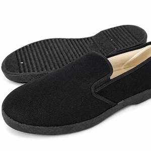 May include: Black slip-on shoe with a textured sole, positioned in front of a matching sole. The shoe features a rounded toe and a low-profile design. The interior lining is a light beige color.
