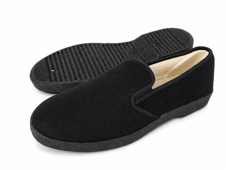 May include: Black slip-on shoe with a textured sole, positioned in front of a matching sole. The shoe features a rounded toe and a low-profile design. The interior lining is a light beige color.
