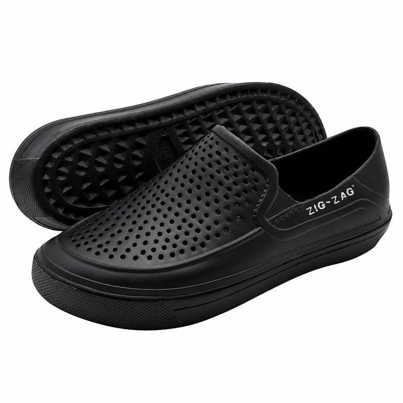 May include: Black slip-on shoes with a perforated upper design. The shoes have a rounded toe and a thick, textured sole. The brand name "ZIG-ZAG" is printed on the side of the shoe. One shoe is positioned on top of the other.