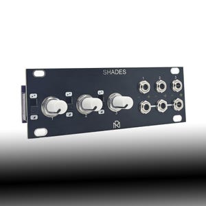 Op de afbeelding: Een donkergrijze elektronische module met het opschrift "SHADES", witte knoppen en zilveren aansluitingen. De module heeft drie grote knoppen en diverse kleinere aansluitingen en poorten. De module is ontworpen voor audio- of elektronische toepassingen.
