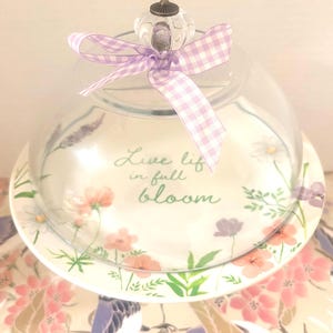 May include: A clear glass cake dome with a floral design and a lavender gingham ribbon. The plate features a floral pattern with the words "Live life in full bloom". The dome has a glass knob on top.