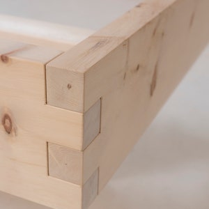 K&ouml;nnte beinhalten: Nahaufnahme einer Holzeckverbindung, die die komplizierte Schwalbenschwanzkonstruktion zeigt. Das Holz ist hellbraun und hat eine nat&uuml;rliche Maserung.