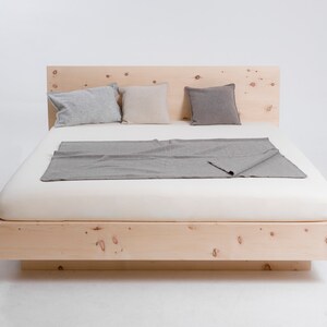 K&ouml;nnte beinhalten: Ein Holzbettgestell mit naturbelassenem Holzfinish. Das Bett hat ein Kopfteil und ein Fu&szlig;teil und ist aus massivem Holz gefertigt. Das Bett ist nicht bezogen, und auf dem Kopfteil liegen drei Kissen.