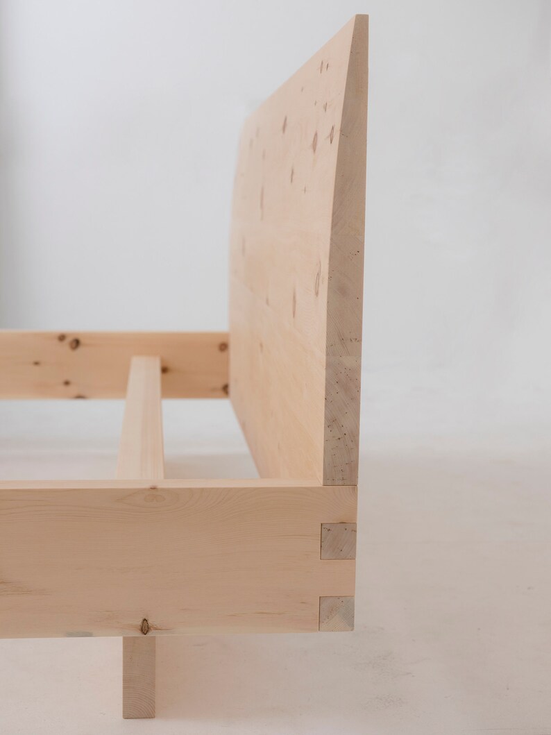 K&ouml;nnte beinhalten: Ein Holzbettgestell mit einem Kopfteil aus einem einzigen St&uuml;ck Holz. Das Kopfteil ist oben abgerundet und hat eine nat&uuml;rliche Holzoptik. Der Rahmen ist aus massivem Holz gefertigt und hat ein schlichtes Design.