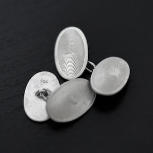 May include: A pair of oval silver cufflinks featuring a concentric circular pattern on the face. One cufflink shows the hallmark 935 stamped on the back. The metal has a matte finish and the design consists of fine, etched lines radiating outward.