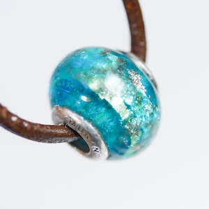 May include: A round, teal and blue bead with silver accents and a brown leather cord. The bead has a marbled appearance with silver flecks. The silver ring around the bead has etched text. The cord is a dark brown color.