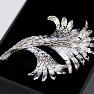 May include: A silver brooch with a floral design, displayed in a black box. The brooch has a detailed, textured surface and a flowing, organic shape. The brooch is a decorative accessory.