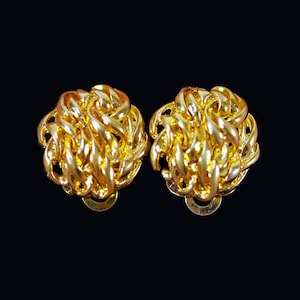 May include: A pair of gold-colored clip-on earrings. Each earring features a woven, rounded design, creating a textured, knotted appearance. The earrings are set against a solid black background, highlighting their metallic sheen.