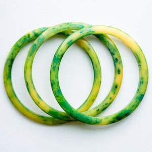 May include: Three circular bangles featuring a marbled pattern of translucent yellow and vibrant green. The bracelets have a smooth, rounded profile and are arranged in an overlapping formation, showcasing the blended color variations throughout.