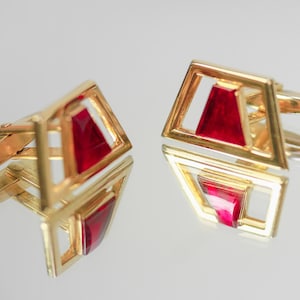 May include: A pair of gold-coloured cufflinks with a geometric design. Each cufflink has a red, triangular centre within a square frame. The cufflinks are displayed on a reflective surface.