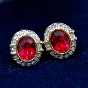 May include: A pair of oval stud earrings. Each earring has a bright red gemstone encircled by small, clear crystals. The setting includes gold-coloured accents and a silver-coloured metal base, set against a dark blue background.