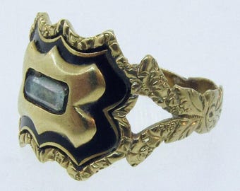 May include: A gold ring with a rectangular central stone. The ring has a shield-shaped face with black enamel details and floral patterns on the band. The ring's design is vintage.