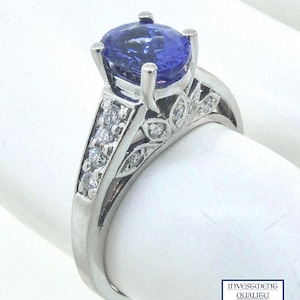 Puede incluir: Un anillo plateado con una gran piedra preciosa azul de corte ovalado como pieza central. La banda presenta piedras preciosas más pequeñas y transparentes, y recortes decorativos en forma de hojas. El anillo está sobre un fondo blanco. El texto "INVESTMENT QUALITY JEWELS" es visible.