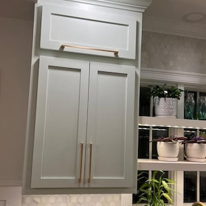 May include: A set of light gray kitchen cabinets featuring flat-panel doors and a single upper cabinet door, all accented with long, slender, metallic copper-toned horizontal and vertical bar handles for a clean, modern appearance.