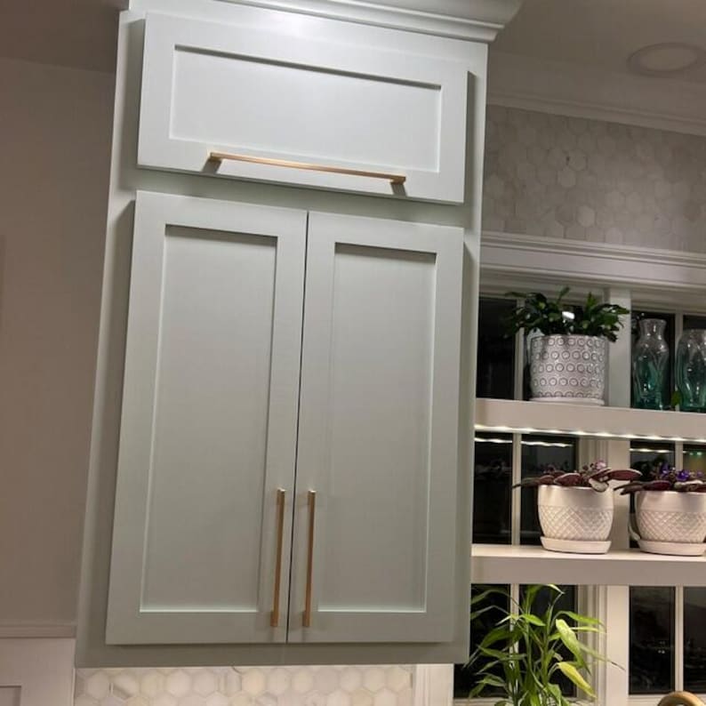May include: A set of light gray kitchen cabinets featuring flat-panel doors and a single upper cabinet door, all accented with long, slender, metallic copper-toned horizontal and vertical bar handles for a clean, modern appearance.
