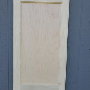 May include: A plain, unfinished wood cabinet door with a raised panel. The door is light brown and has a simple, rectangular frame.