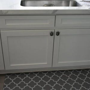 May include: White kitchen cabinets featuring dark metal turtle-shaped cabinet knobs. The cabinets have a flat-panel design with a rectangular frame detail. The hardware is mounted on the upper corner of each cabinet door.