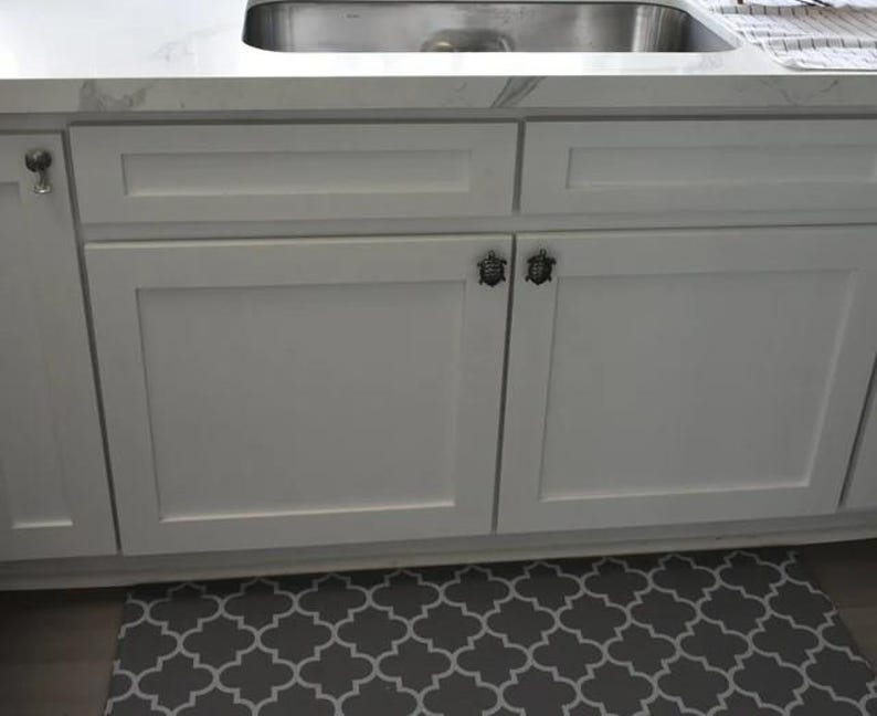 May include: White kitchen cabinets featuring dark metal turtle-shaped cabinet knobs. The cabinets have a flat-panel design with a rectangular frame detail. The hardware is mounted on the upper corner of each cabinet door.