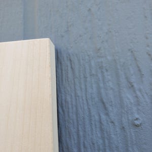 May include: A rectangular piece of light-colored, smooth-grained wood board with a clean, straight edge. The wood features a natural, pale beige tone with subtle grain patterns visible across its flat surface and a sharp, defined corner.