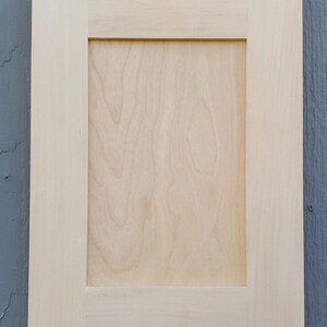 May include: A rectangular cabinet door featuring a light-colored, unfinished wood finish with a visible natural grain pattern. The design consists of a flat center panel surrounded by a sturdy frame with clean, straight edges and corners.