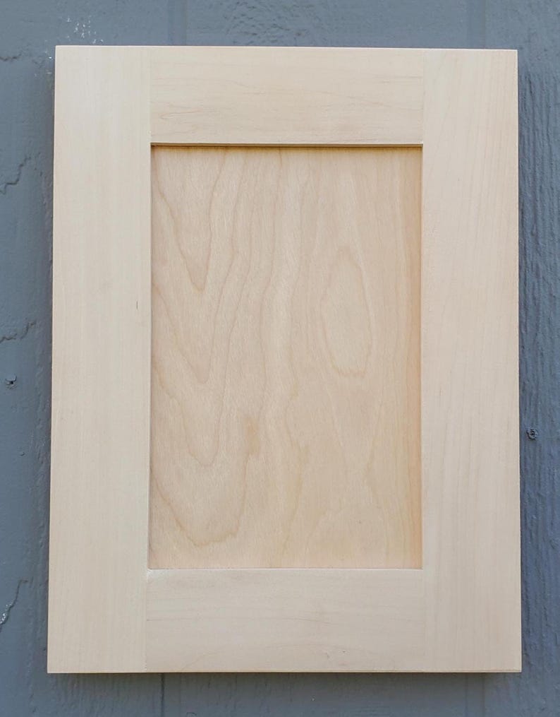 May include: A rectangular cabinet door featuring a light-colored, unfinished wood finish with a visible natural grain pattern. The design consists of a flat center panel surrounded by a sturdy frame with clean, straight edges and corners.