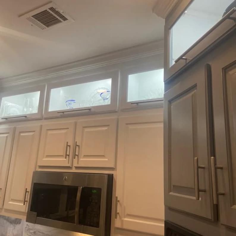 May include: White kitchen cabinetry featuring upper glass-front cabinets with interior lighting, silver metal handles, and a stainless steel microwave oven integrated into the lower section of the wall-mounted storage units.