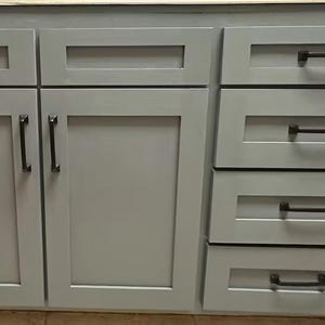 May include: A set of light gray kitchen cabinets featuring shaker-style doors and drawers. Each cabinet door and drawer is equipped with a dark bronze metal handle. The cabinetry has a smooth, matte finish and a clean, rectangular design structure.