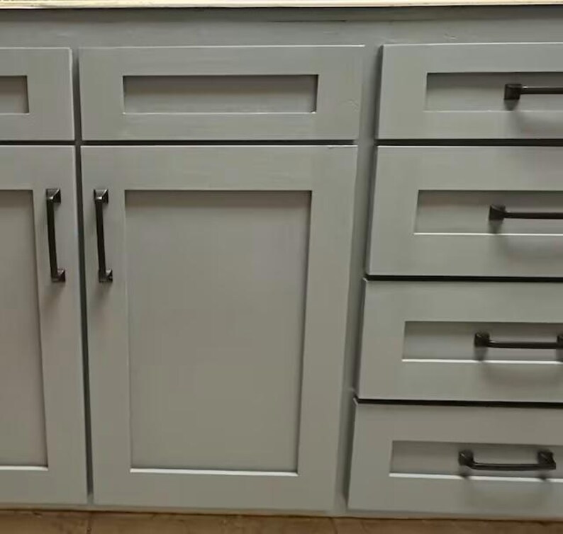 May include: A set of light gray kitchen cabinets featuring shaker-style doors and drawers. Each cabinet door and drawer is equipped with a dark bronze metal handle. The cabinetry has a smooth, matte finish and a clean, rectangular design structure.