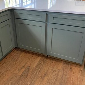 May include: Sage green kitchen cabinetry featuring a shaker-style design with flat recessed center panels and horizontal drawer fronts. The cabinets are topped with a smooth white countertop and installed above a light brown wood floor.